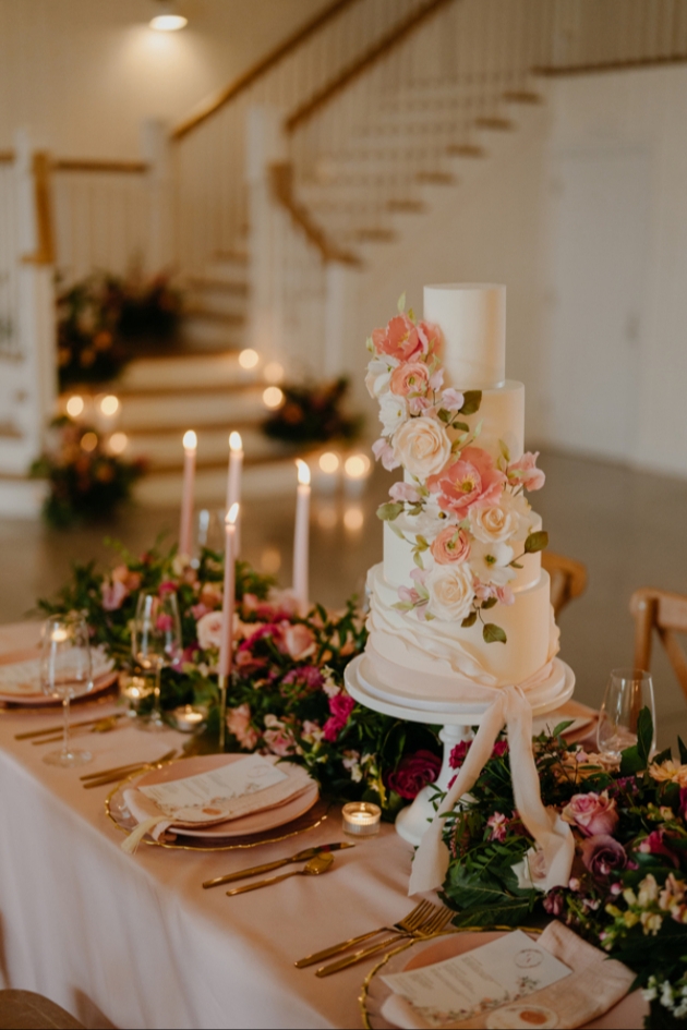 intimate wedding tablescape in peach featuring ornate wedding cake adorned in sugar flowers, nestled among pink floral table runner.