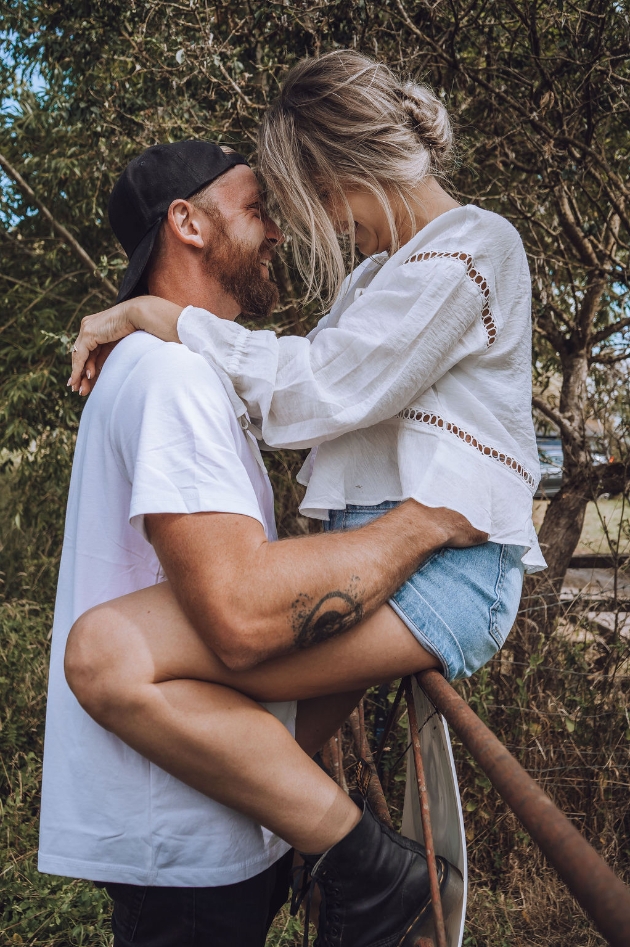 Engaged couple woman wears denim shorts and sits on a metal gate man wears white t-shirt and black baseball cap
