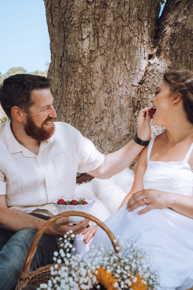 Engaged couple - man feeds the woman strawberries as the sit beneath a tree with picnic