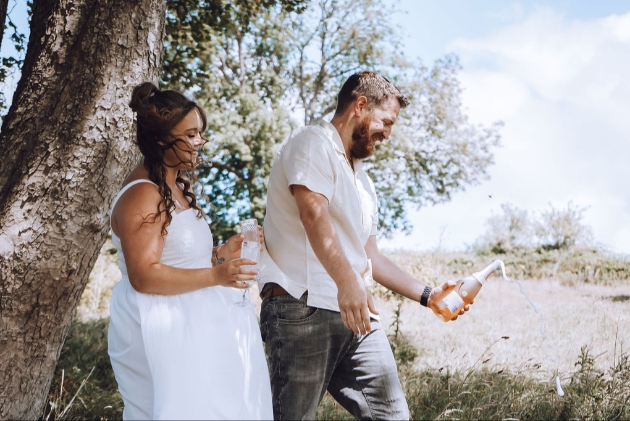 Engaged couple - man opens bottle of bubbly while woman holds champagne glass - countryside setting