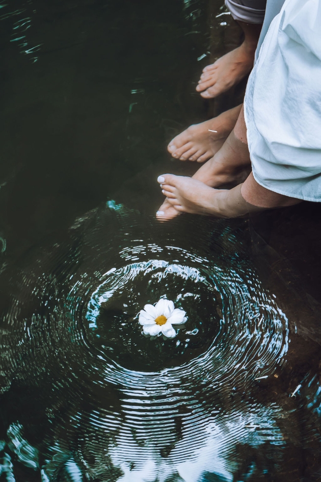 Engaged couple dangled their feet over a river above a white daisy they've dropped in